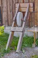 An antique grindstone standing against a wooden shed with overgrown grass. Old grey sharpening stone tool or equipment used outside on a farm