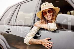 One beautiful caucasian woman hanging out the window of a car while taking a roadtrip. Attractive young female hipster with tattoos on her arm, wearing a hat and sunglasses and taking a drive in a car