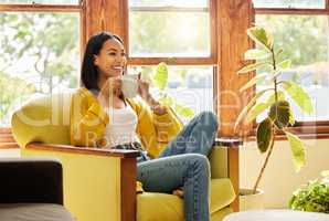 Woman relaxing, drinking coffee sitting in front of a window in a bright living room. A happy smiling young hispanic female looking past the camera sitting on a chair enjoying her break on a chair at home
