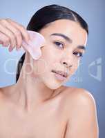 Studio portrait of a beautiful mixed race woman using a rose quartz gua sha for deep penetration cell renewal. Young hispanic woman using anti ageing tool against blue copyspace background