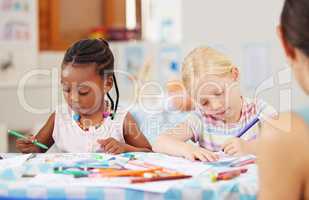 African american girl sitting at a table and colouring at pre-school or kindergarten with her caucasian classmate . Young female children using colourful pencils to draw pictures in a class at school