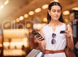 A young mixed race woman using a cell phone and bank card while carrying bags during a shopping spree. Young brunette woman purchasing items online with her smartphone and credit card in a shopping mall. Woman using banking app to increase spending limit