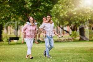 Mixed race family in the park. Happy young mother and father piggybacking their son and daughter outside. Couple carrying their kids while walking on grass during. Two sisters on parents backs