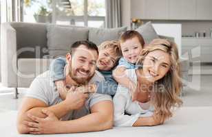 Portrait of smiling caucasian family of four relaxing on floor in lounge at home. Playful sons lying and clinging on carefree loving parents backs while bonding and spending fun quality time together