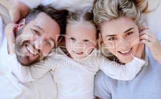 Portrait of happy carefree caucasian family in pyjamas from above lying cosy together in bed at home. Loving parents with little daughter. Cute young girl touching and hugging her mom and dads face