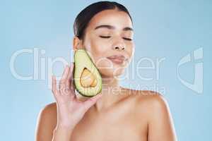 Beautiful young mixed race woman with an avocado isolated in studio against a blue background. Her skincare regime keeps her fresh. For glowing skin, eat healthy. Packed with vitamins and nutrients