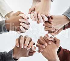 Below close up view of people holding hands in circle shape. A group of people putting their hands together while standing in a huddle inside against a clear grey background. Anything is possible with teamwork