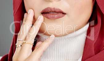 Muslim woman wearing rings, lipstick, a hijab, touching her face and posing in studio. Closeup of half headshot of a confident arab model isolated against grey background. Fashionable middle eastern