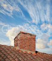 A brick chimney on the roof in the blue cloudy sky background. A closeup of the chimney on the rooftop. An orange bricked roof with chimney against the blue sky with copy space on a sunny day.
