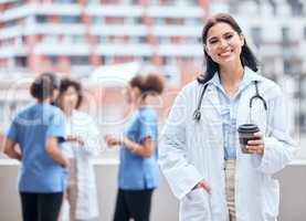 Portrait of smiling young doctor enjoying a cup of coffee outside with her colleagues in the background. Happy medical professional taking a break from working in the hospital to drink tea.