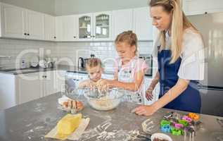 Caucasian mother and little daughters baking together in a kitchen at home. Mom teaching girls how to make dough in a messy kitchen. Sisters learning how to bake with their mom