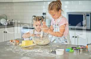 Two messy little girls baking in the kitchen at home. Caucasian focused sisters learning to make a dessert and cook. Adorable children bonding and helping each other to prepare a recipe for the house