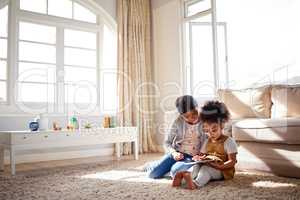 Two cute little mixed race sisters sitting on the carpet and playing games on digital tablet together at home. Adorable hispanic female friends learning to use a wireless device together