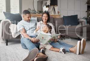 A happy mixed race family of three relaxing on the lounge floor and reading a story book at home. Loving black family being affectionate on a carpet. Young couple bonding with their son at home
