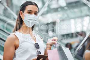 Portrait of a young mixed race woman wearing a medical face mask for prevention against coronavirus while holding a cellphone and shopping bags. Fashionable hispanic carrying retail bags after buying in a mall during Covid-19 pandemic