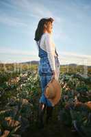 Woman farmer standing in a cabbage field on a farm. Young female with a straw hat and rubber boots looking over her field of organic vegetables