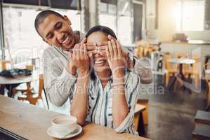 Cheerful young man covering his girlfriends eyes and surprising her while sitting in a cafe. Happy young mixed race couple meeting for coffee on their first date. Excited woman trying to guess who is behind her