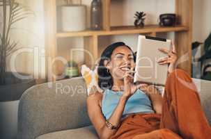 Smiling mixed race woman browsing internet on digital tablet at home. Happy hispanic lying down on living room sofa alone and using technology. Relaxed woman scrolling and searching online on weekend