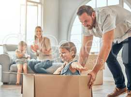 A happy mature caucasian father pushing his daughter in a box while her mother and sister sit on the sofa in the lounge at home. Man and girl having fun, playing games at home while enjoying family bonding. Family looking overjoyed in their new home