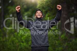 Young hispanic fit male athlete cheering with his fists in the air while on a run in a forest outside in nature. Exercise is good for health and wellbeing. Happy to reach his fitness goals