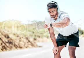 Athletic young mixed race man listening to music and looking at his smartwatch while exercising outdoors. Handsome hispanic resting while checking the time during a workout. Tracking his progress