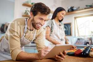 Close up of happy young man browsing on digital tablet searching for recipe to follow online while cooking at home with his wife