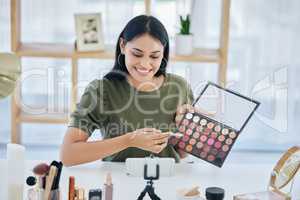 One beautiful mixed race creative woman sitting alone in a studio and using her cellphone to vlog while showing her eyeshadow palette. Happy hispanic makeup artist filming a tutorial for the internet
