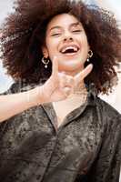 Portrait of smiling beautiful trendy mixed race woman with afro making cool hand gesture to pose alone outside. Below view of fashionable hispanic showing rock emoji. African American feeling playful