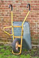 Garden wheelbarrow leaning against a red brick wall in a home backyard. Landscaping equipment and tools to carry and transport soil, manure, compost and fertilizer. Done cleaning a landscaped yard