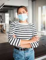 Young mixed race businesswoman standing with her arms crossed and wearing a mask at work. One hispanic female boss wearing a mask protecting from a virus and standing at work