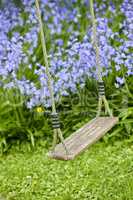 A wooden swing in a peaceful garden area with Bluebell Flowers blooming in a lush green bush. A play space outdoors in a backyard with vibrant purple plants growing in on a spring day
