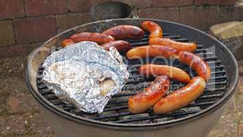 Food stuff. Sizzling barbecue sausages and chicken wrapped in foil outdoors in a yard on a hot day. Delicious meat roast on a grill for a cookout by a home or house hosting a weekend party.