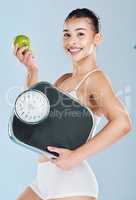 Portrait of a happy young woman holding an apple and scale against blue copyspace background. Confident athletic fit female showing the benefits of a balanced diet, weight loss and healthy lifestyle