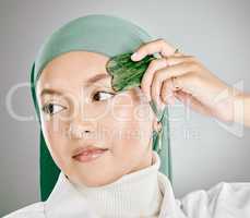 A gua sha being used on a young muslim womans face, isolated against grey studio background. Woman wearing a hijab or headscarf, using an anti ageing tool to reduce wrinkles and promote cell renewal