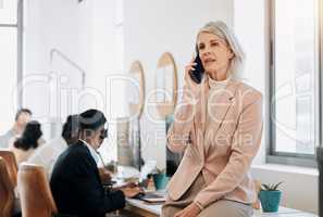 What can I do for you. a mature businesswoman sitting in the office and using her cellphone while her team work behind her.