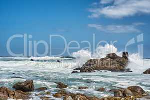 Big waves splashing and breaking on the coastline. Turbulent sea with rough tides from strong winds crashing onto beach boulders with a blue sky background. Rocky coast in Camps Bay, South Africa