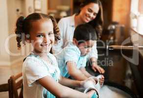 Happy cheerful little mixed race girl helping her mother wash the dishes in the kitchen at home. Hispanic child smiling while washing a cup with soap and water. Family keeping their house clean