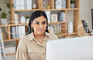 Young indian businesswoman working on a computer in an office. One female only browsing online while planning on a desktop pc at her desk. Confident hardworking entrepreneur completing deadlines