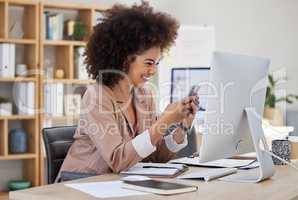 Happy young mixed race businesswoman with afro texting on cellphone while working on computer in an office. One female only checking social media and browsing apps online on smartphone to manage plans