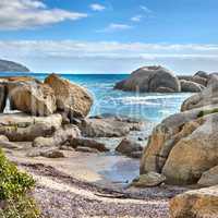 Landscape of beautiful large boulders in the ocean with a blue cloudy sky. Rock or granite structures shining under the sun near calm foamy waves at a popular beach location, Cape Town, South Africa