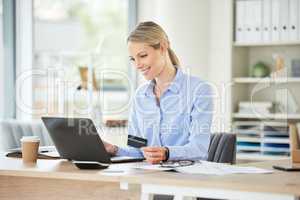 Beautiful young caucasian woman reading her credit card while using her phone and laptop to shop online while sitting in the office at work. Shopping has never been simpler or more convenient