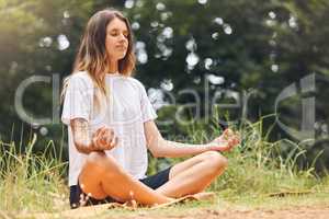 A calm young caucasian female sitting with her legs crossed doing yoga and meditating on grass in a park on a sunny day. Young woman focusing her zen and energy while working on her mental wellness and relaxing in a forest