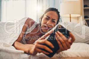 A young mixed race woman looking annoyed while scrolling social media and lying in bed. An attractive Hispanic female using her cellphone and getting bad news while resting in her bedroom