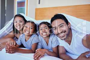 Happy mixed race family of four in pyjamas lying cosy together in a row on a bed with blanket over their heads at home. Loving parents with two kids. Adorable girls bonding with mom and dad at bedtime