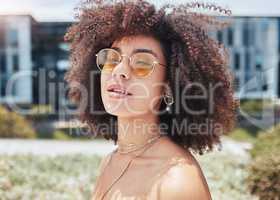 Cool, trendy african american woman with a curly afro wearing sunglasses outside. Face of a young serious mixed race woman enjoying a summer day while standing outside. Hipster girl in the city