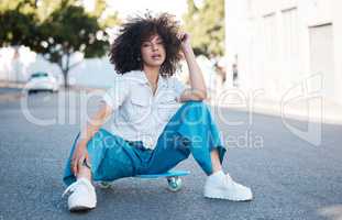 A young female mixed race woman skate sitting on a skateboard looking cool and confident with great style in a street outside. Hispanic hipster woman with curly afro hair style in a cool outfit against a urban background