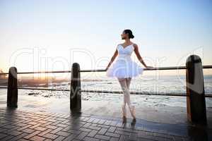 Elegant young ballerina wearing white fancy dress and pumps while leaning against a railing on the promenade with a beautiful ocean background at sunset
