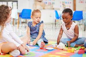 Group of diverse adorable little girls sitting cross legged on the floor in preschool and having a tea party. Three cute children playing and bonding in school. African American kid with her friends
