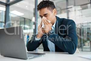 Sick mixed race businessman blowing his nose with a tissue while working on a laptop alone at work. One hispanic male businessperson suffering from allergies blowing his nose in an office