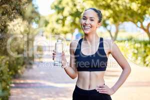 Portrait of one fit young mixed race woman taking a rest break to drink water from bottle while exercising outdoors. Happy female athlete quenching thirst and cooling down after running and training workout